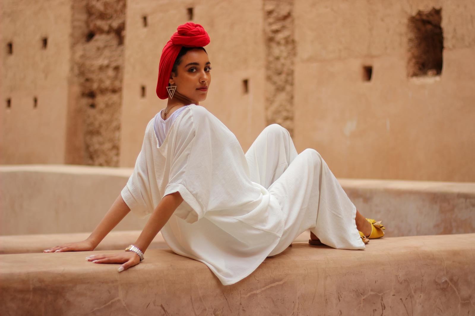 Stylish woman in white outfit with red headscarf poses outdoors in Marrakesh courtyard.Morocco Dress Code