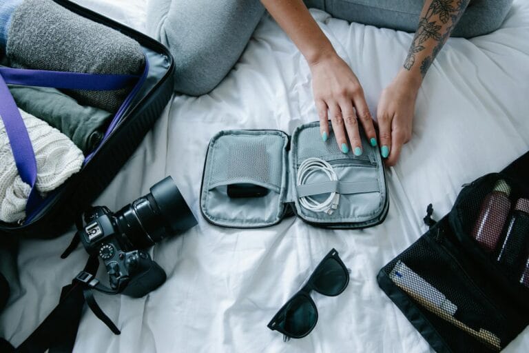 Woman with tattoos packs electronics, camera, and sunglasses in a suitcase on a bed.What to wear in Morocco