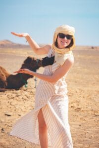 A woman in a striped dress and headscarf smiling with a camel in the Moroccan desert.Merzouga Weather in December