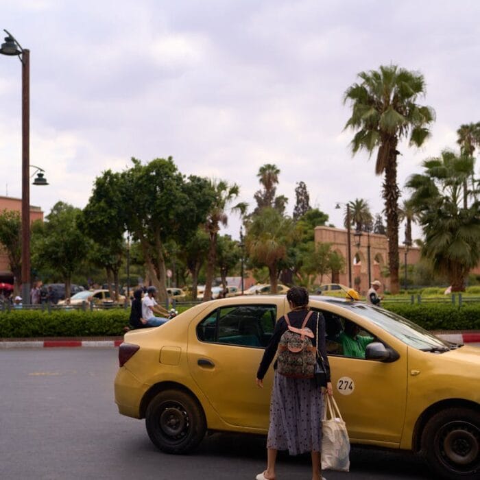 A woman enters a yellow taxi on a street in Marrakesh, surrounded by palm trees and city life.How to get a Taxi in Marrakech