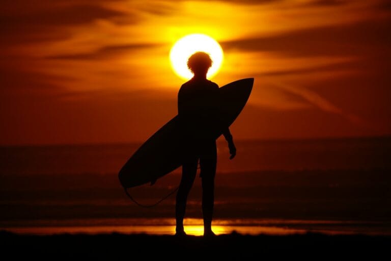 A silhouette of a person holding a surfboard at sunset.Family Surfing Vacations Morocco