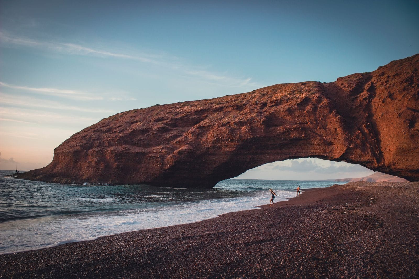 Breathtaking view of the natural arch at Lagzira Beach, Morocco, during sunset with people enjoying the shoreline.beach destinations morocco