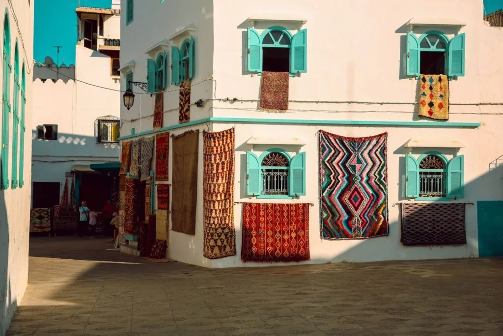 Vibrant street in Asilah, Morocco featuring traditional woven rugs hung on white buildings with turquoise shutters.Is Asilah Worth Visiting for Families