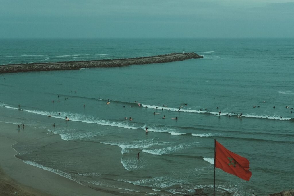 A tranquil beach with waves, Moroccan flag, and surfers enjoying the oceanWhen Is the Best Time to Visit Morocco’s Beaches.