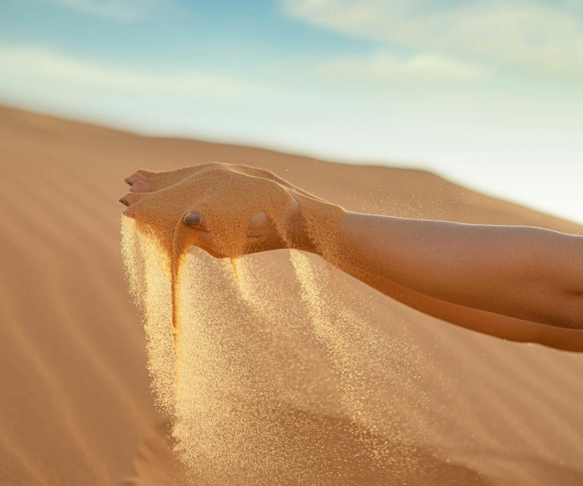 A hand releasing fine sand against the vast landscape of Merzouga Sand Dunes, Morocco.errachidia to merzouga