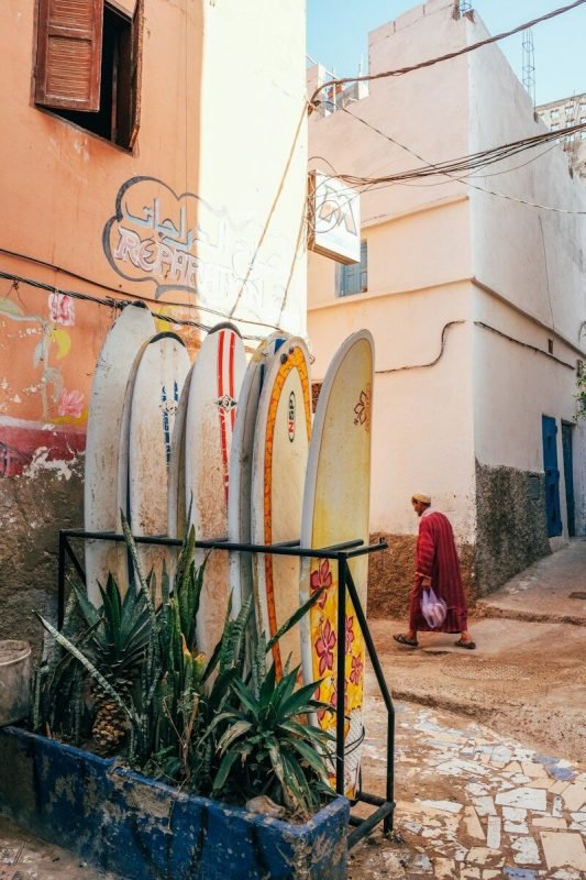 Surfboards lean against a sunlit building, with a man walking by